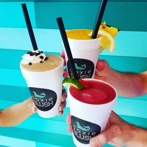 Three colorful slush drinks in white cups with straws, topped with whipped cream and fruit, against a teal background.
