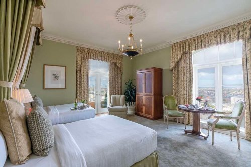 Luxurious hotel room with two beds, elegant decor, a chandelier, and a view of the city through large windows.