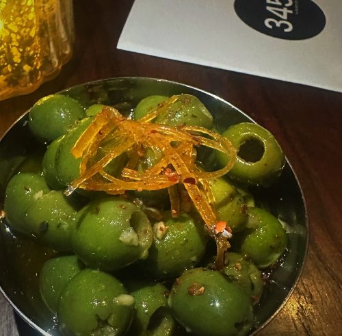 A bowl of green olives topped with thin strips of orange zest and spices, set on a wooden table.