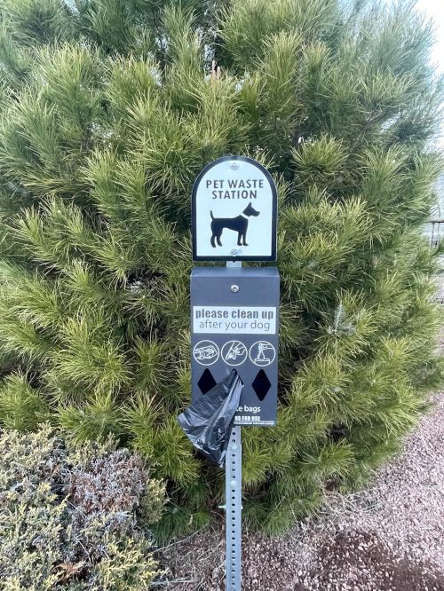 Pet waste station with a sign and bag dispenser, surrounded by greenery. Instructions to clean up after dogs.