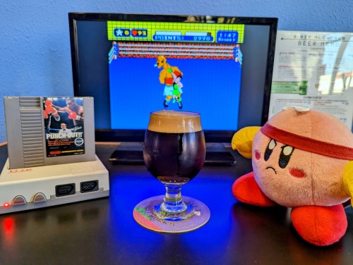 A retro gaming setup with a NES console, a glass of dark beer, and a plush Kirby next to a boxing game on screen.