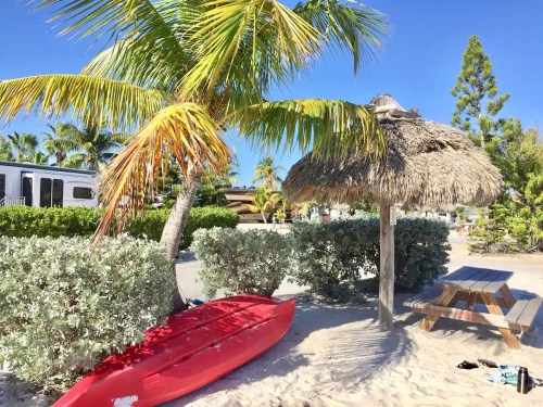 A red kayak rests on sandy beach near a thatched hut and palm trees under a clear blue sky.