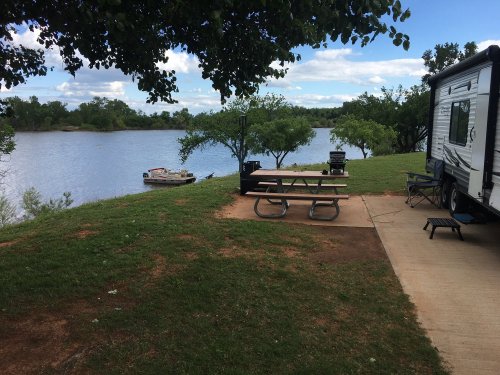 A serene lakeside scene featuring a picnic table, RV, and a small boat on the water, surrounded by greenery.