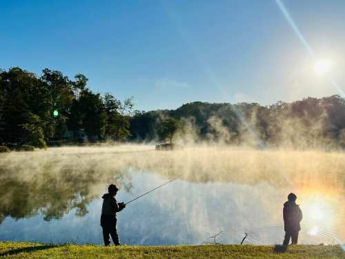 Two people fishing by a misty lake at sunrise, surrounded by trees and a calm, reflective water surface.