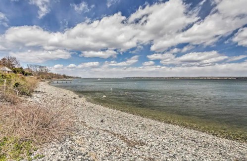 A rocky shoreline with calm water under a partly cloudy sky, featuring distant land and a few boats.