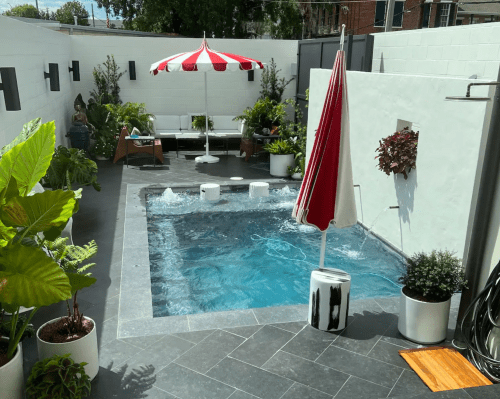 A modern backyard with a small pool, red and white umbrella, lounge chairs, and lush greenery.