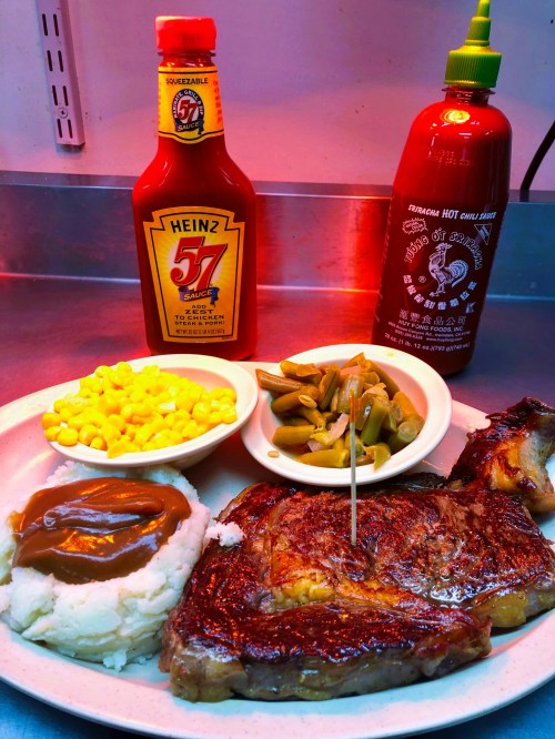 A plate with a grilled steak, mashed potatoes, green beans, corn, and sauces in the background.