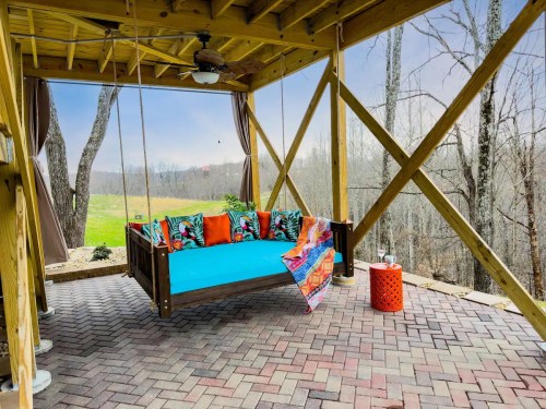 A colorful swing bed with cushions under a wooden structure, surrounded by trees and a grassy landscape.