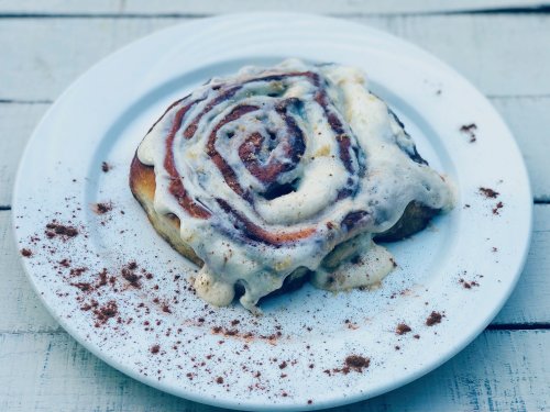 A delicious cinnamon roll topped with creamy icing, served on a white plate with a sprinkle of cinnamon.
