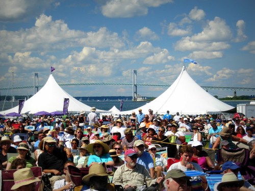 A large crowd sits under tents at an outdoor event, with a bridge and blue sky in the background.