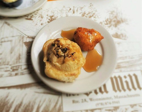 A plate with a sweet roll topped with nuts and a fried dough ball, drizzled with syrup, on a patterned tablecloth.
