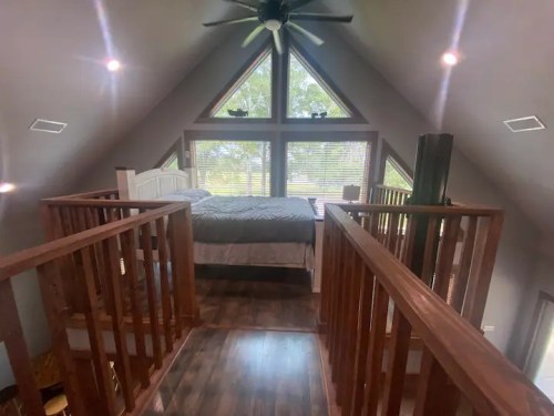 Cozy loft bedroom with a triangular window, wooden railings, and a bed, surrounded by natural light.
