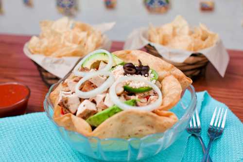 A colorful taco salad in a crispy bowl, topped with chicken, onions, green peppers, and olives, with tortilla chips in the background.