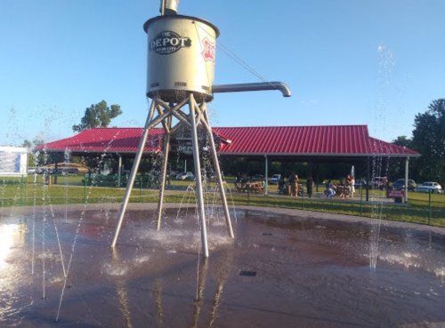 A large water tower structure with a sign reading "The Depot" stands over a splash pad, with a red-roofed building in the background.