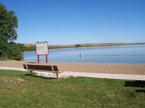A serene lakeside view with a bench, sandy shore, and a sign, surrounded by green grass and distant hills under a clear blue sky.