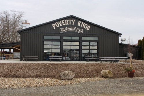 Exterior of Poverty Knob Farmhouse Ales building, featuring large windows and a gravel area with benches.