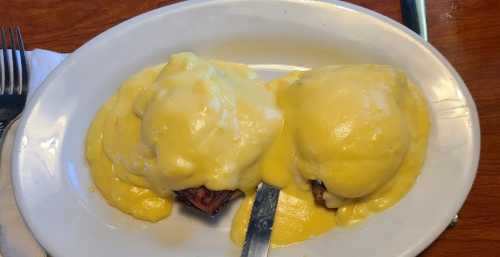 Two eggs Benedict on a white plate, topped with creamy hollandaise sauce, served with a knife.