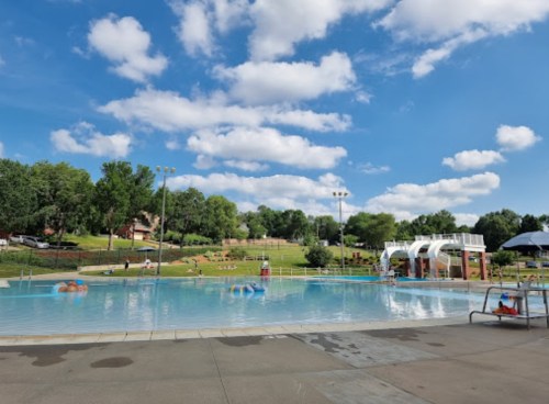 A sunny day at a swimming pool with clear water, lounge chairs, and green trees in the background under a blue sky.