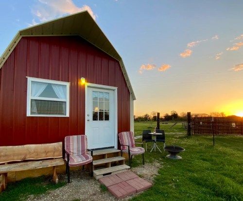 A small red cabin with a white door, surrounded by grass, chairs, and a fire pit, against a sunset backdrop.