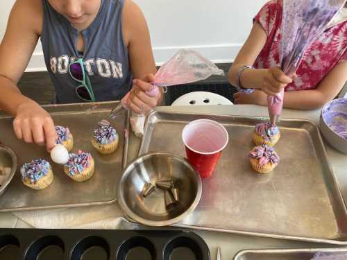 Two children decorate cupcakes with colorful frosting and sprinkles, using piping bags and a spoon.