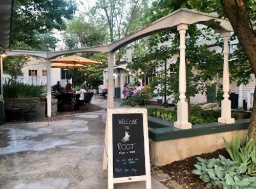 A cozy outdoor dining area with a welcome sign for "Root" surrounded by greenery and patrons enjoying their meals.