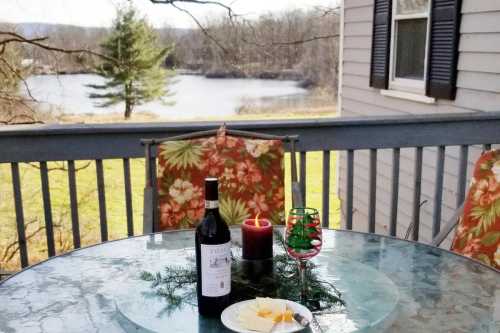 A cozy outdoor table with a bottle of wine, cheese, and a candle, overlooking a serene lake and trees.