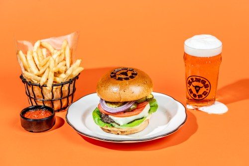 A burger with lettuce, tomato, and onion, served with fries and a pint of beer on an orange background.