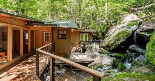 A wooden cabin nestled in a lush forest, with a small waterfall and rocky landscape nearby.