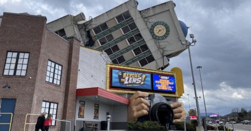 A tilted building with a large camera sculpture in front, advertising "Beyond the Lens!" under a cloudy sky.