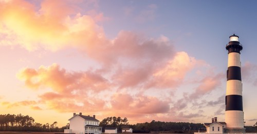 A black and white striped lighthouse stands beside a white house under a colorful sunset sky with fluffy clouds.
