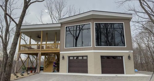 Modern two-story house with large windows, a wooden deck, and a gravel driveway, surrounded by bare trees.