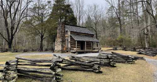 A rustic log cabin surrounded by a wooden fence, set in a wooded area with bare trees and a gravel path.