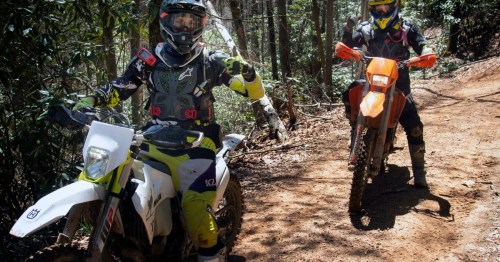 Two motocross riders on dirt bikes, giving a thumbs up on a forest trail. Sunlight filters through the trees.