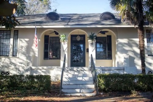 A charming house with a front staircase, arched windows, and an American flag, surrounded by greenery.
