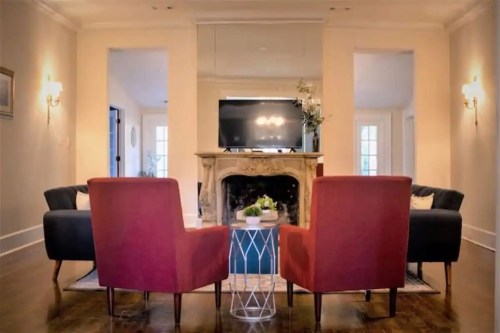 A cozy living room with two red chairs, a blue sofa, a coffee table, and a TV above a fireplace. Bright and inviting space.