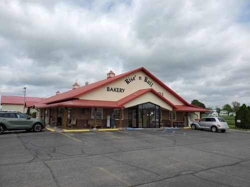 A bakery building with a red roof and sign reading "Rise 'n Roll Bakery," surrounded by parked cars and cloudy skies.