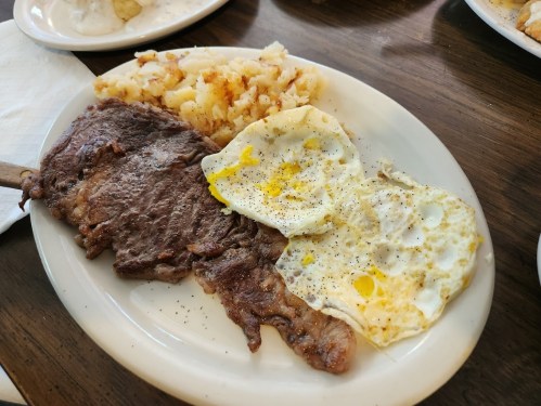 A plate with a grilled steak, two fried eggs, and crispy hash browns on a wooden table.