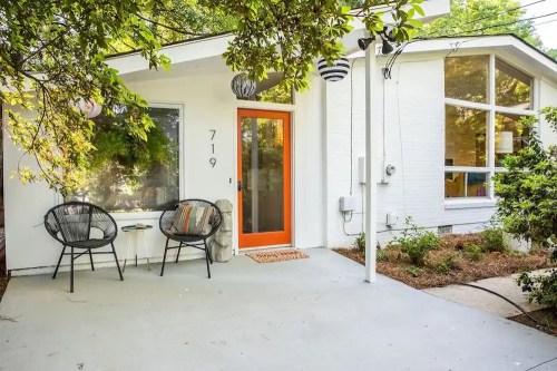 A modern white house with an orange door, featuring a small porch and two black chairs under a tree.