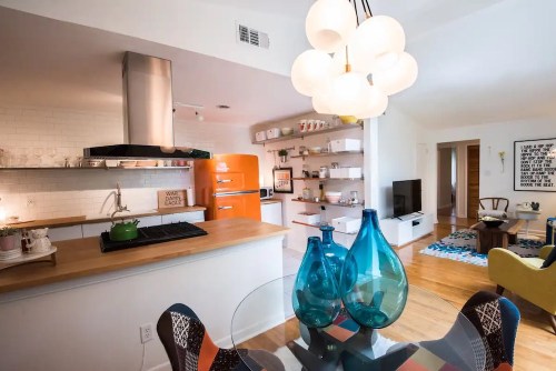 A modern kitchen with an orange fridge, wooden shelves, and a dining table featuring blue vases and colorful chairs.