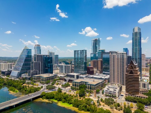 Aerial view of a vibrant city skyline with modern skyscrapers and a river, under a clear blue sky with fluffy clouds.