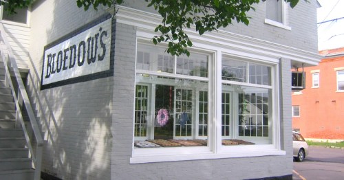 A quaint bakery with large windows displaying various baked goods, surrounded by greenery and a brick building.