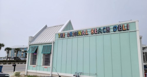 A light blue building with a sign that reads "The Original Beach Deli," set against a cloudy sky.