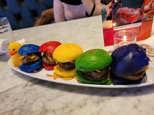 A platter of colorful mini burgers in red, yellow, green, blue, and purple buns, served with a candle and a rubber duck.