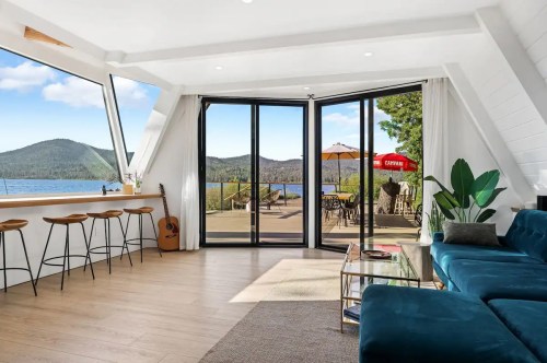 Bright, modern living space with large windows, lake view, and outdoor seating area. Cozy seating and wooden bar stools.