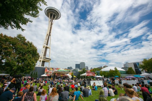 A crowd gathers on a grassy area with the Space Needle in the background, enjoying an outdoor event under a cloudy sky.
