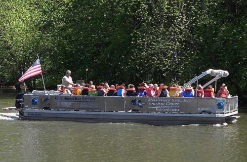 A tour boat with passengers wearing life jackets cruises on a river, surrounded by lush greenery and an American flag.