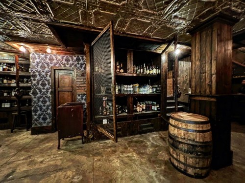 A rustic bar interior featuring wooden shelves, a barrel, and a vintage decor with patterned wallpaper.