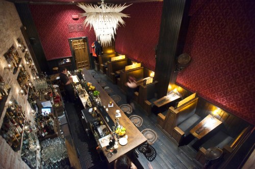 A stylish bar interior with a long counter, wooden seating, and a striking chandelier, featuring warm lighting and rich decor.