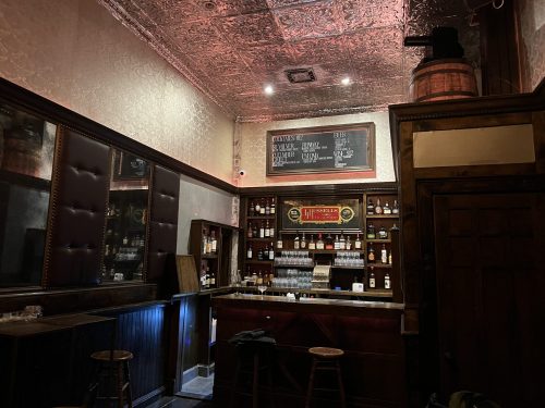 A cozy bar interior with wooden accents, shelves of liquor, and warm lighting, featuring a vintage-style decor.
