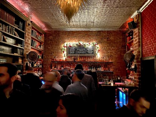 A lively bar scene with patrons mingling, brick walls, shelves of books, and festive decorations.